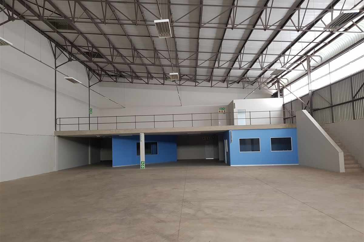 Spacious industrial warehouse featuring a mezzanine level, high steel roof trusses, concrete flooring, and integrated office or storage rooms with blue painted walls.