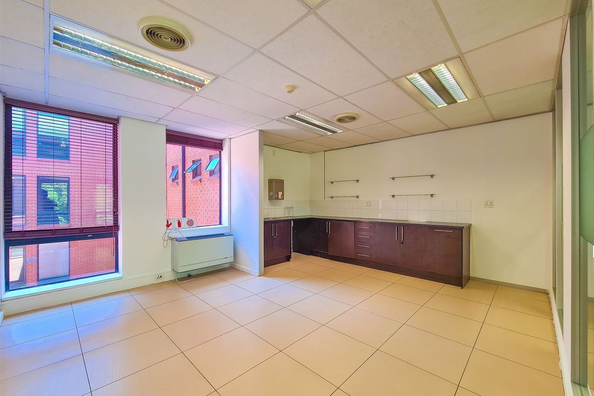 A functional office kitchenette area featuring built-in dark wood cabinetry, tiled flooring, and recessed ceiling lighting with large windows providing natural light.