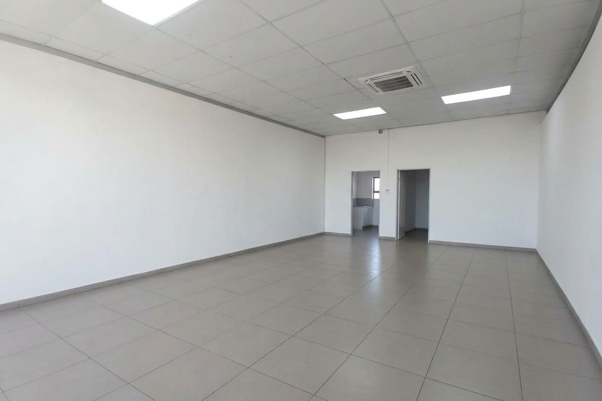 A spacious, empty office area featuring neutral tiled flooring, a suspended ceiling with recessed lighting, and integrated ducted air conditioning.