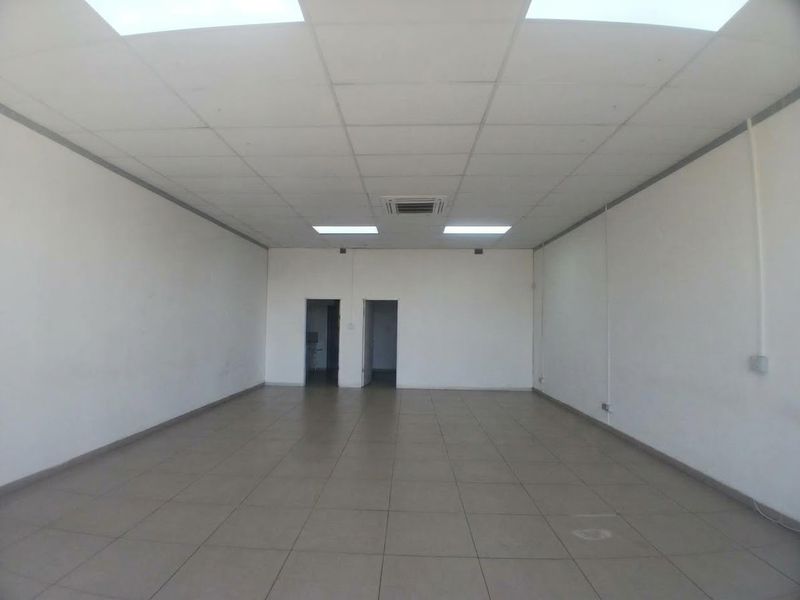 A spacious, empty office area featuring a suspended ceiling, recessed lighting, and neutral tiled flooring, providing a versatile workspace.