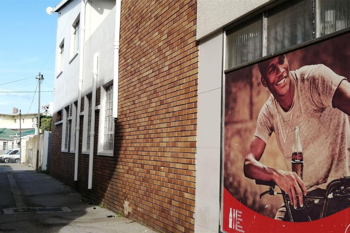 A view of a narrow paved alleyway alongside a commercial building featuring brickwork, downpipes, and a large advertisement on the exterior wall.
