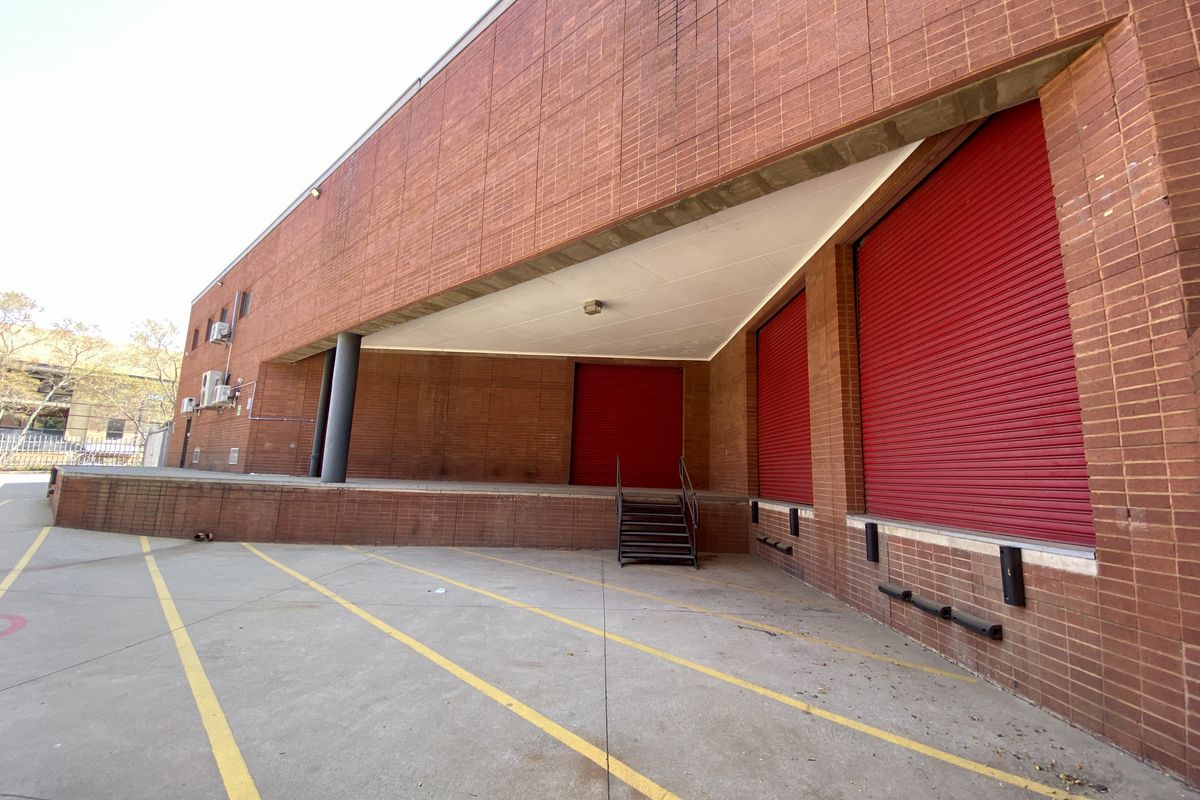 Industrial loading bay area featuring a raised concrete platform, multiple red roller shutter doors, and a covered access point with metal stairs.