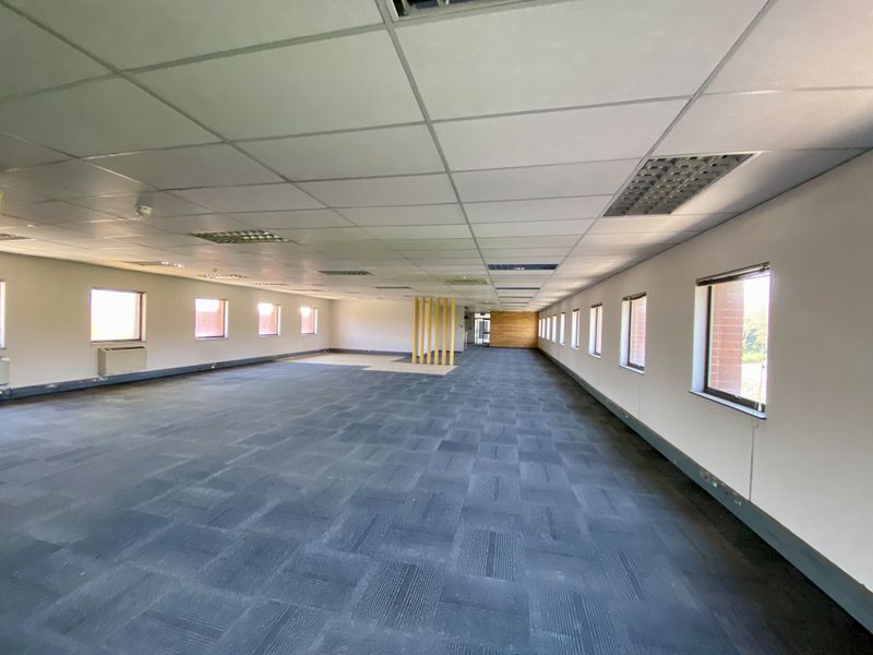A spacious, open-plan industrial office area featuring a suspended ceiling, recessed lighting, and large windows providing ample natural light.