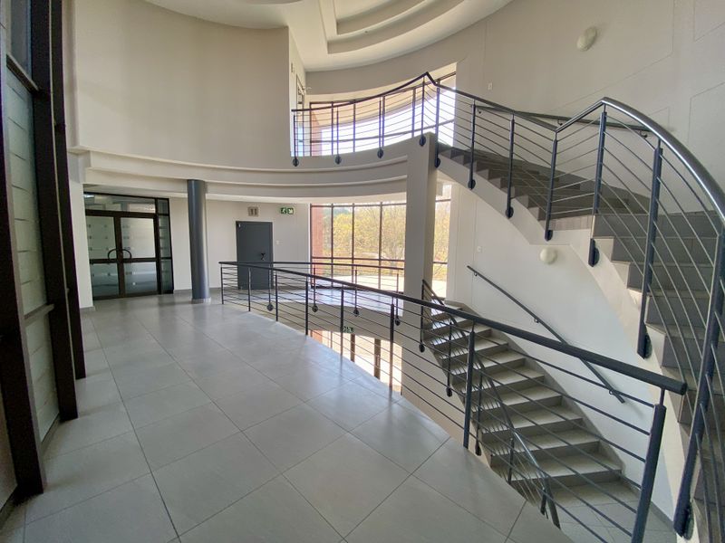 Spacious industrial office building foyer featuring a grand curved staircase with metal railings, tiled flooring, and large glass windows providing natural light.