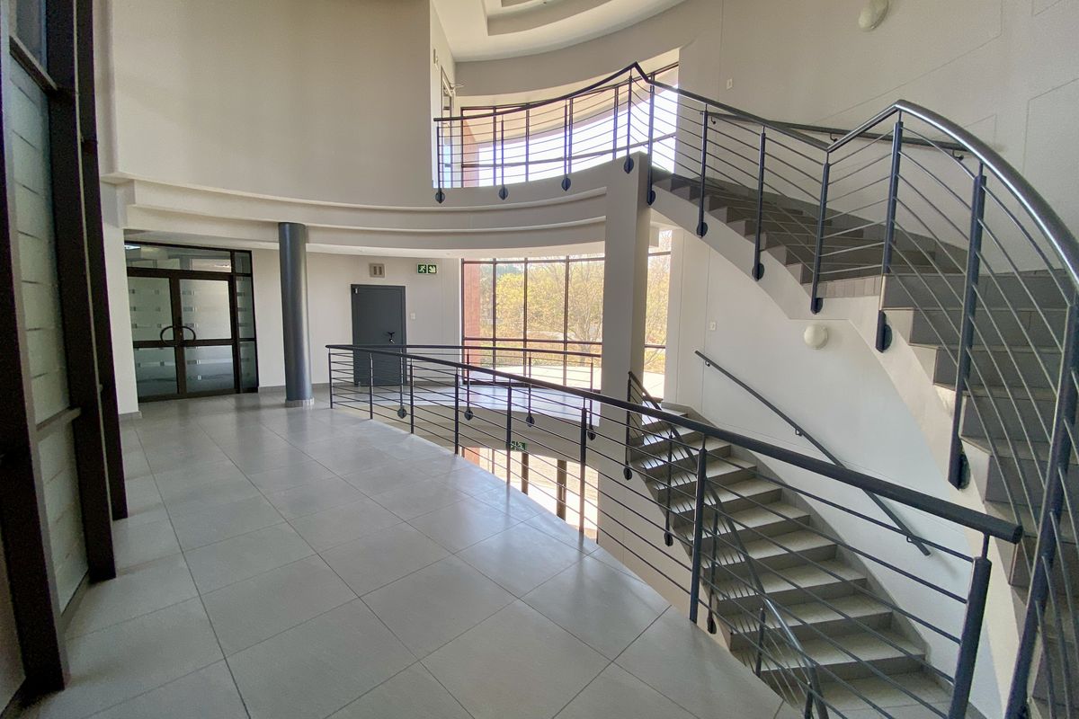 Spacious industrial office building foyer featuring a grand curved staircase with metal railings, tiled flooring, and large glass windows providing natural light.