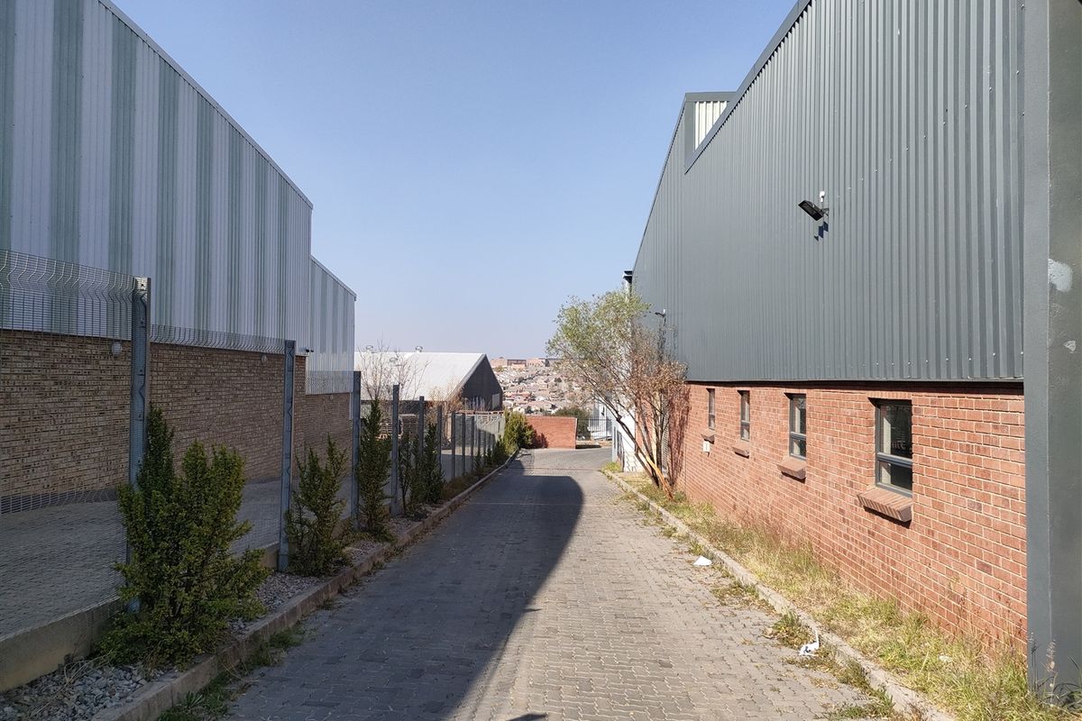 An exterior view of an industrial property featuring a paved access lane between two warehouse buildings with brick and corrugated metal facades.