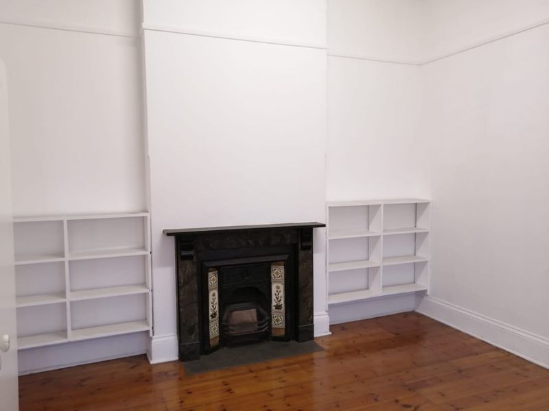 A bright, empty living room featuring polished timber flooring, a classic fireplace with decorative tiling, and symmetrical built-in shelving units.