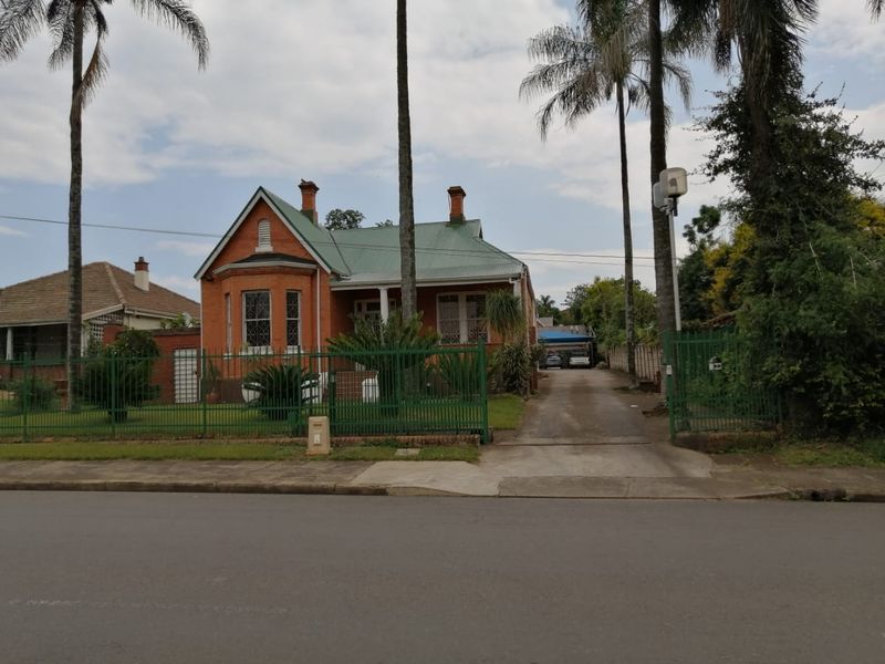 A charming red brick commercial property featuring a corrugated metal roof, leaded glass windows, and a gated concrete driveway with established palm trees.