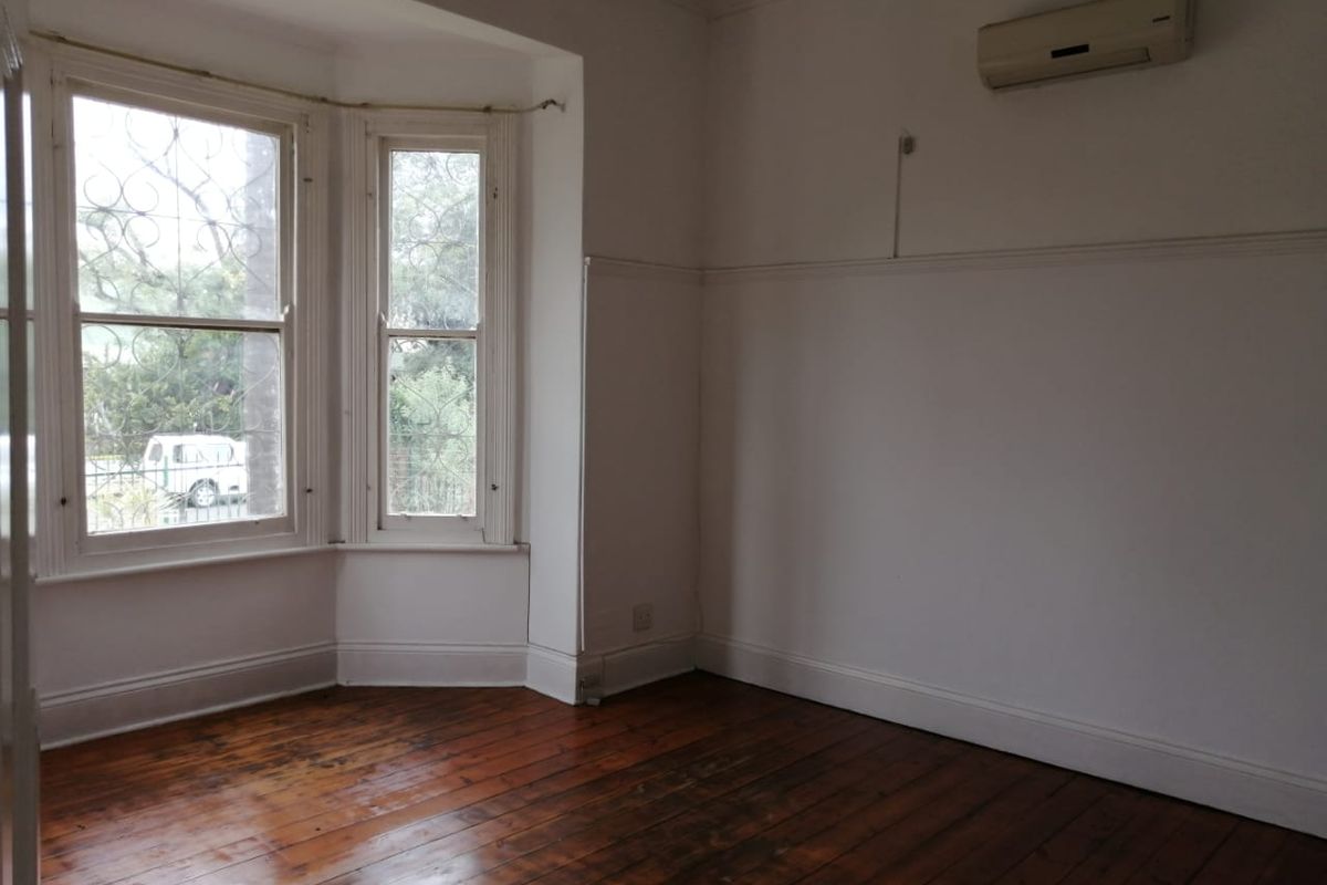 An empty living room featuring original hardwood flooring, a classic bay window with security bars, and a wall-mounted air conditioning unit.
