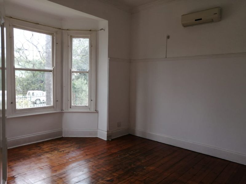 An empty living room featuring original hardwood flooring, a classic bay window with security bars, and a wall-mounted air conditioning unit.