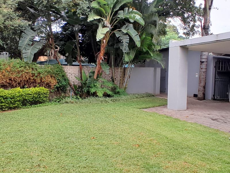 A rear garden area featuring a lawn, established trees, a brick boundary wall, and a covered paved patio or carport structure.