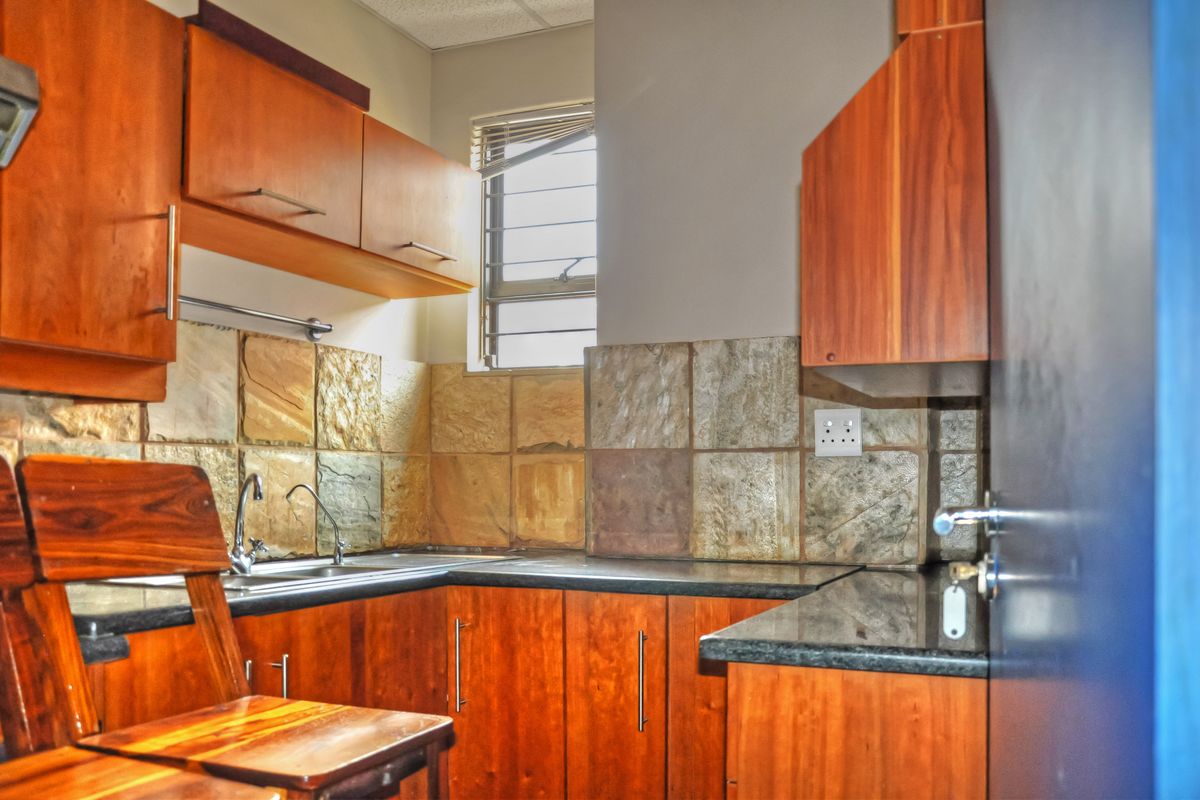 Compact office kitchenette featuring wooden cabinetry, stone-look tile backsplash, dark countertops, and a stainless steel sink.