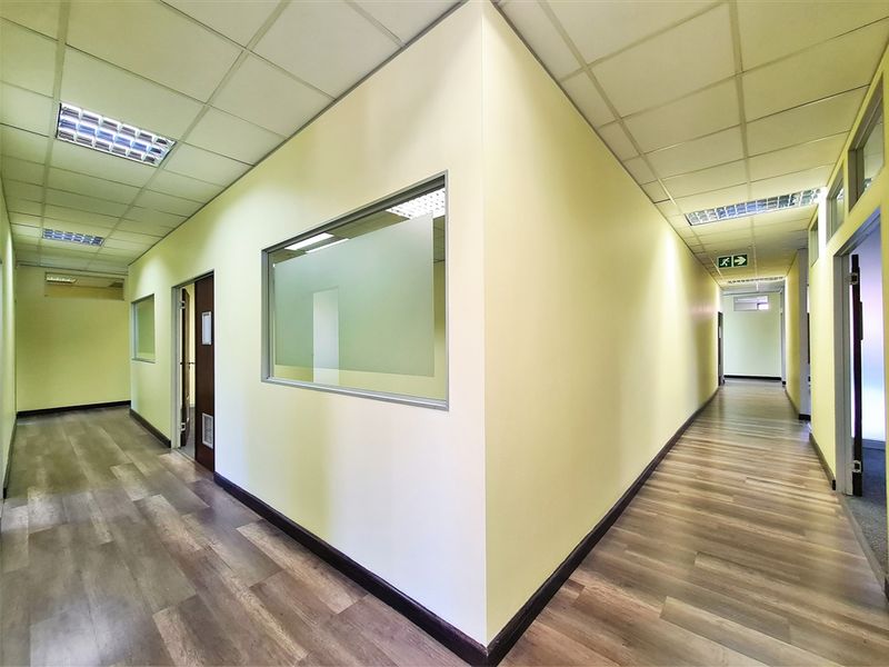 A clean, well-lit office hallway featuring modern laminate flooring, suspended ceilings, and internal glass partitions for a professional workspace environment.