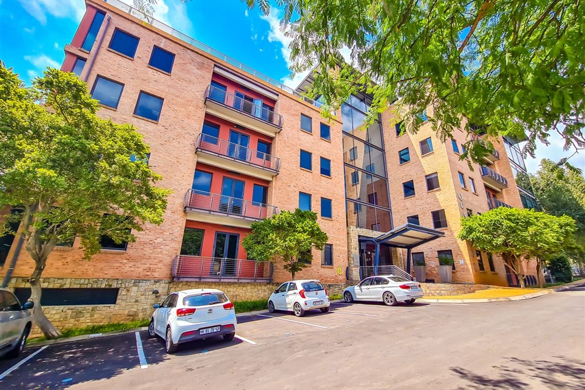 A multi-story office building featuring a brick facade, glass curtain wall, and integrated metal balconies with a paved parking area at the front entrance.