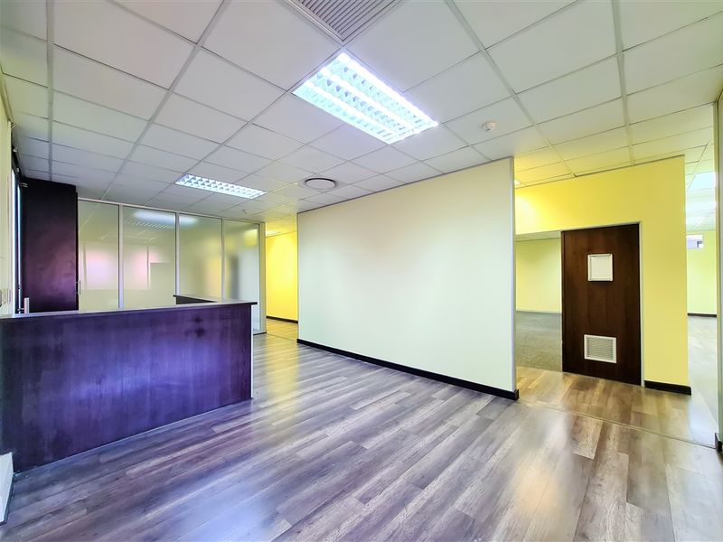 A bright, vacant office space featuring laminate flooring, a built-in reception counter, glass partitions, and a suspended ceiling with recessed lighting.