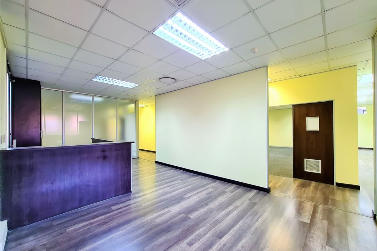 A bright, vacant office space featuring laminate flooring, a built-in reception counter, glass partitions, and a suspended ceiling with recessed lighting.