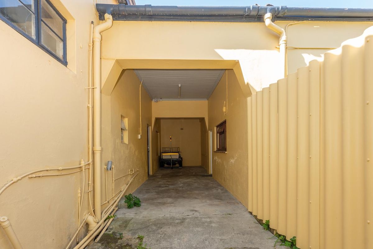 A narrow exterior rear access area featuring a concrete driveway, external plumbing, and corrugated metal fencing, showing signs of wear and weathering.