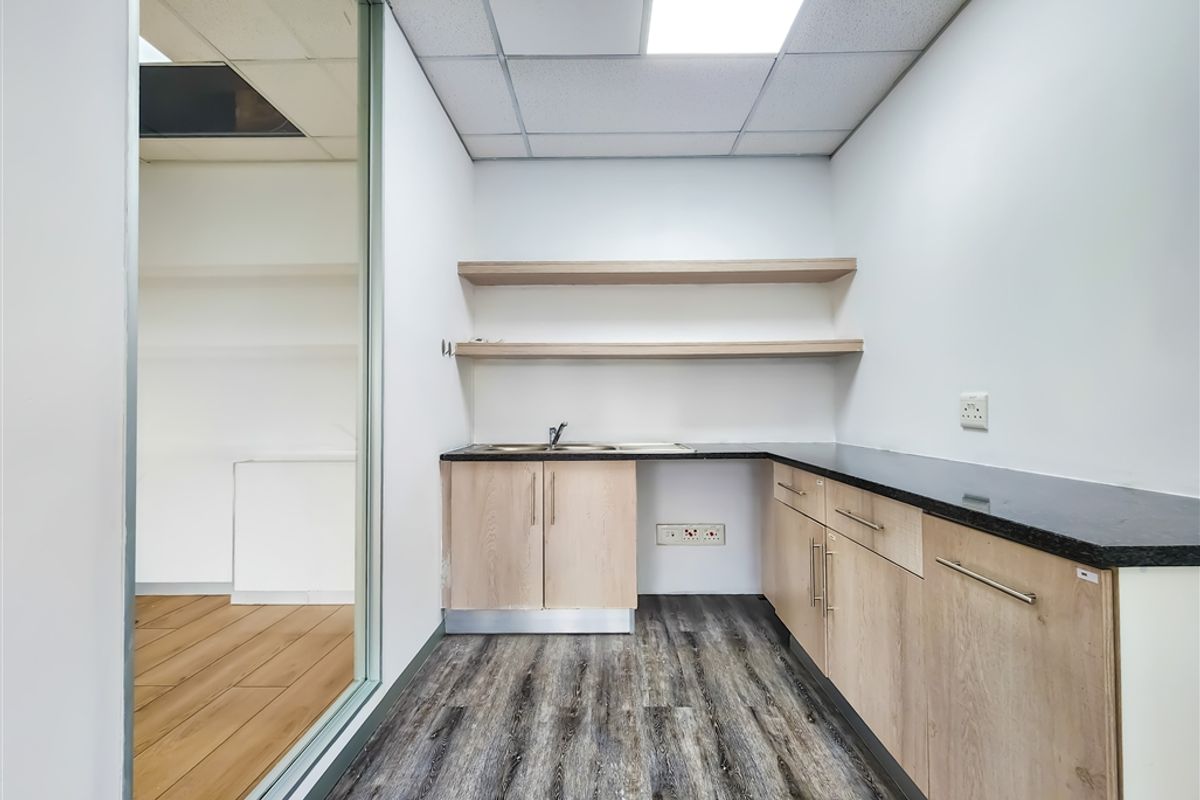 A functional office kitchenette featuring light wood cabinetry, a dark countertop, integrated sink, and open shelving, adjacent to a glass-partitioned office space.