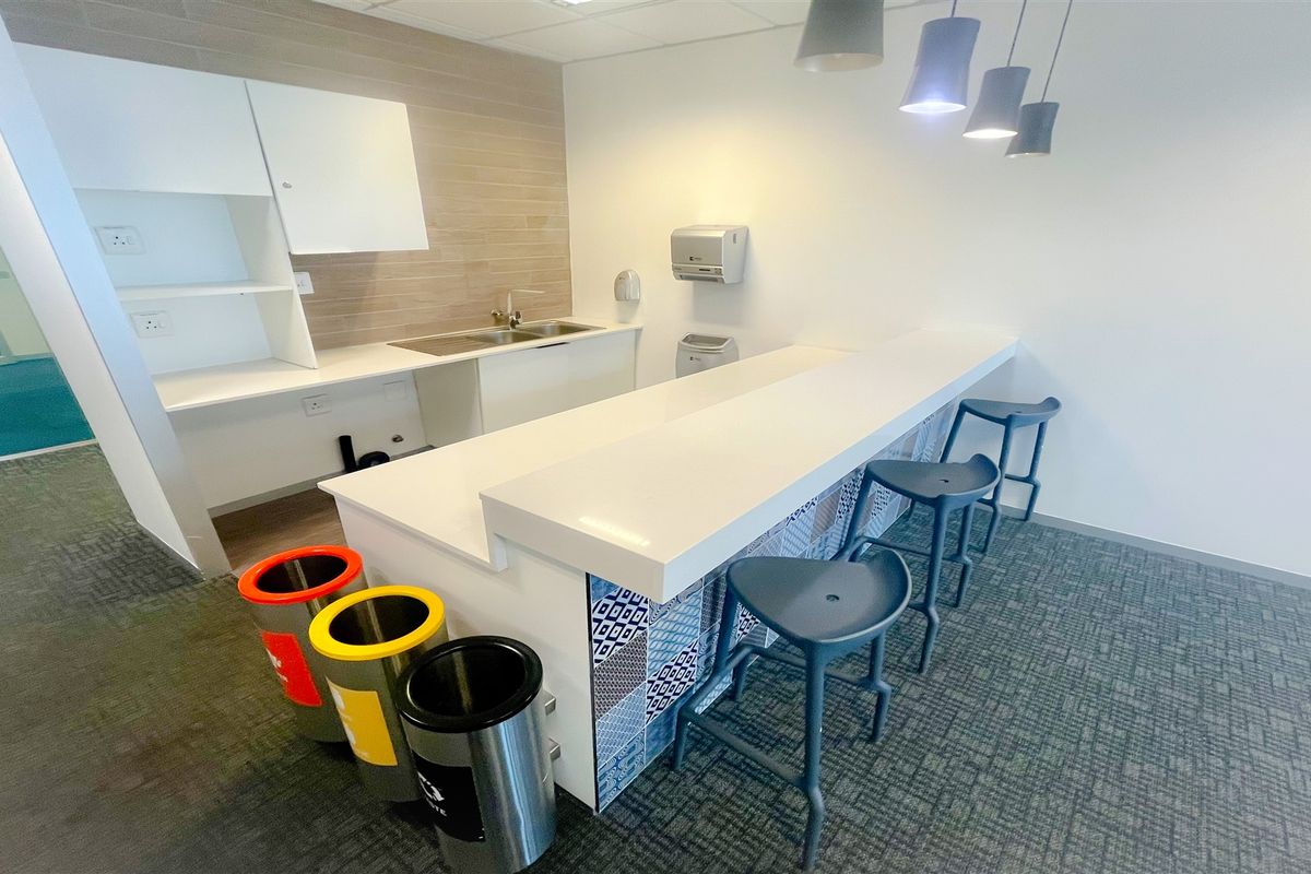 A modern office kitchenette featuring a stone breakfast bar, decorative tiled base, stainless steel sink, and contemporary pendant lighting.