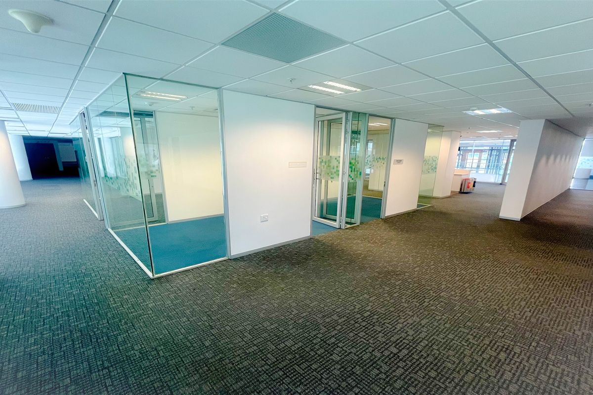 Spacious open-plan office area featuring modern glass-partitioned meeting rooms, suspended ceilings, and durable carpeted flooring throughout.