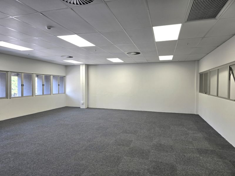 A spacious, empty office area featuring neutral carpet tiles, a suspended ceiling with recessed lighting, and large windows providing natural light.