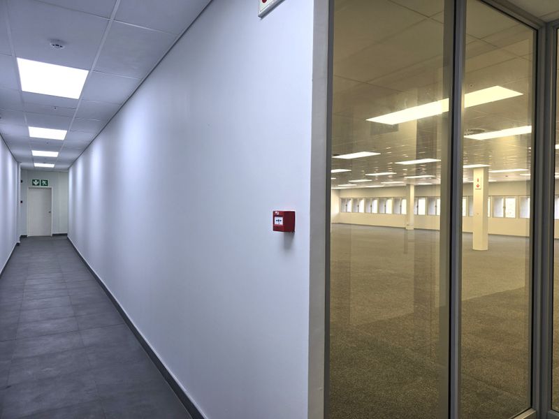 A clean, modern office hallway with tiled flooring and recessed lighting, featuring a glass partition wall looking into an expansive, open-plan office space.