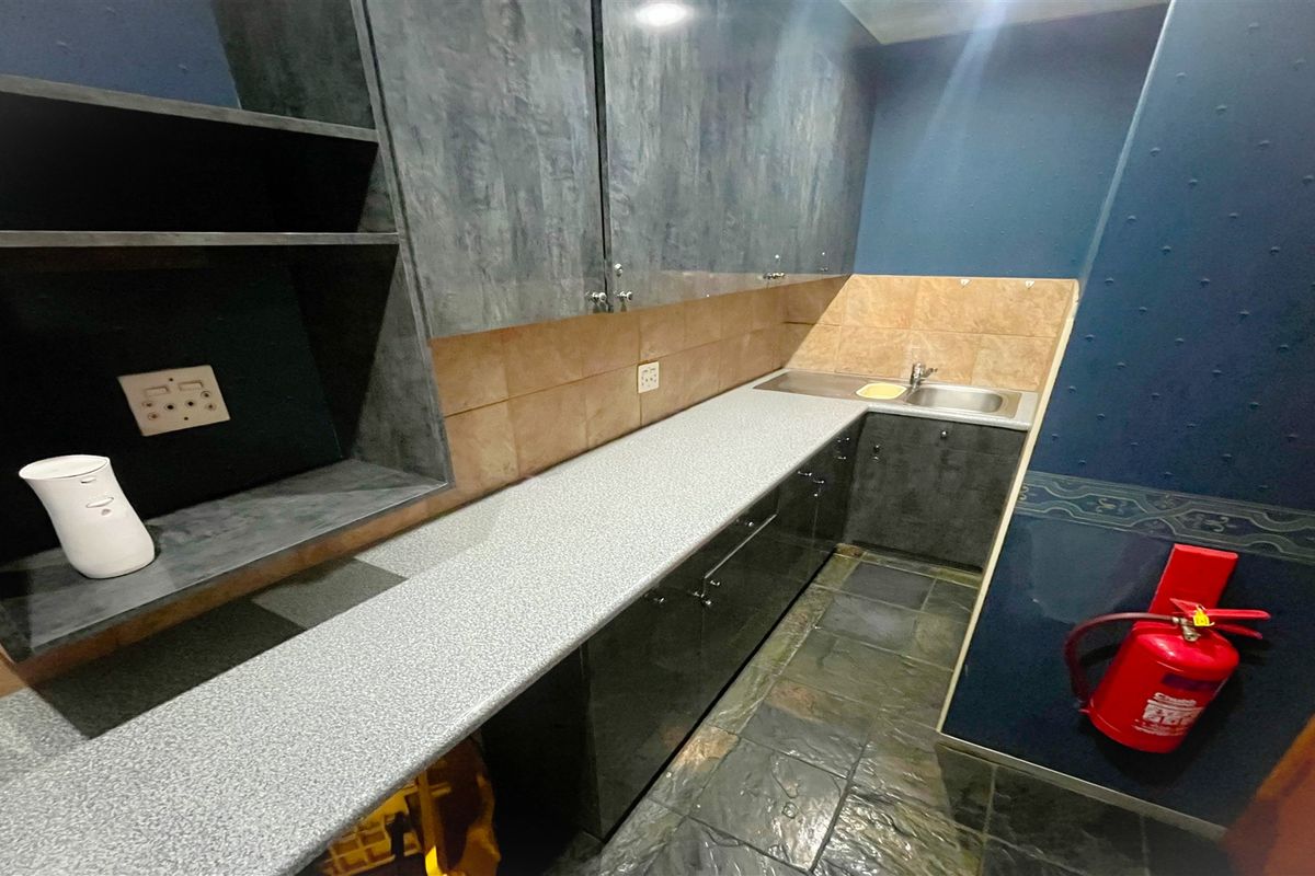 A functional office kitchenette featuring dark cabinetry, a stone-look countertop, stainless steel sink, and tiled backsplash with integrated electrical outlets.