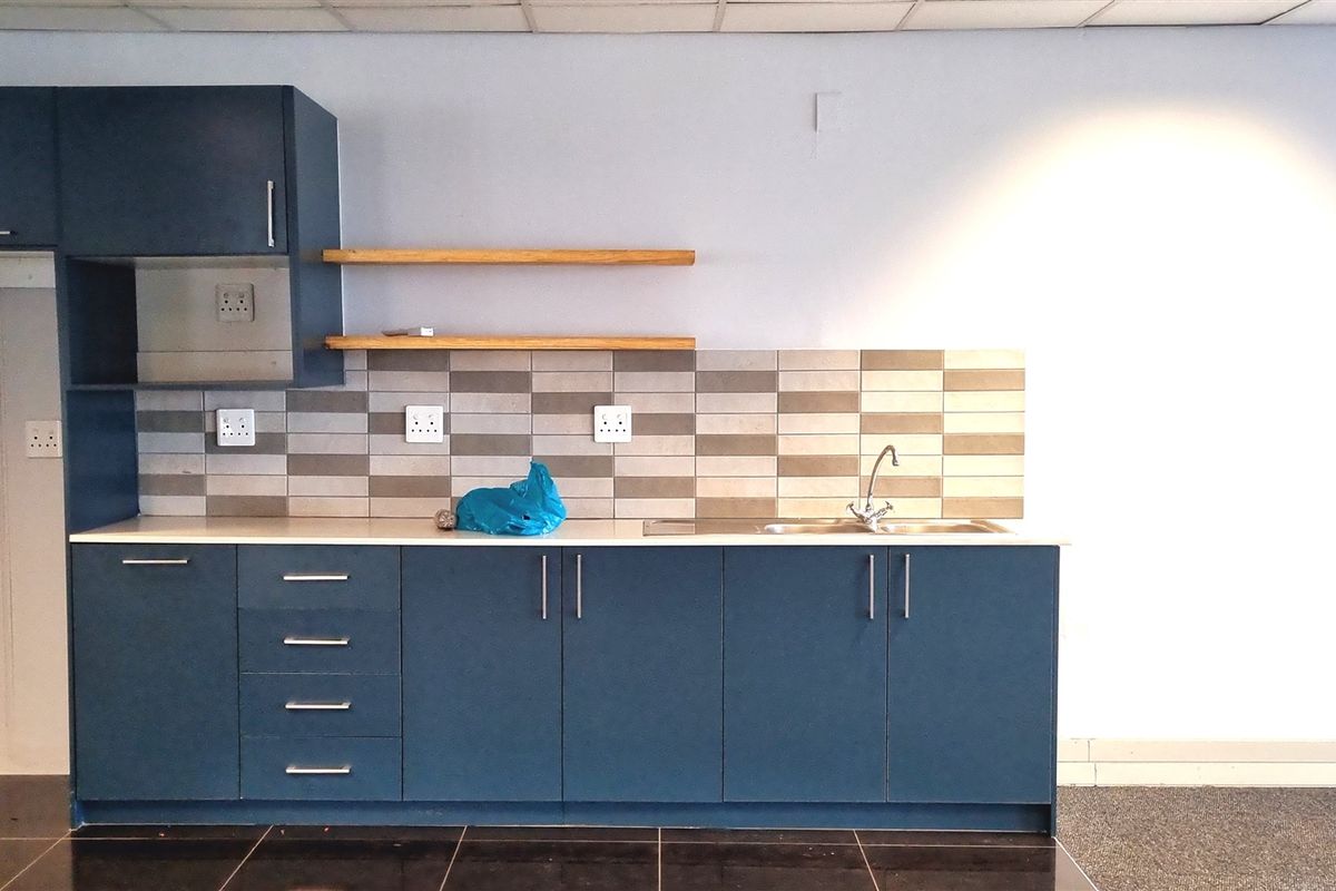 A modern office kitchenette featuring dark blue cabinetry, a stainless steel sink, tiled backsplash, and floating wooden shelves.