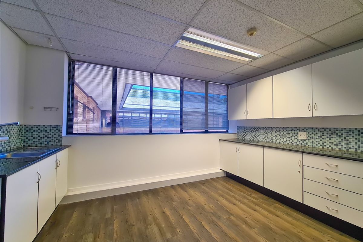 A functional office kitchenette featuring white cabinetry, mosaic tile backsplash, dark countertops, and wood-look flooring with large windows providing natural light.