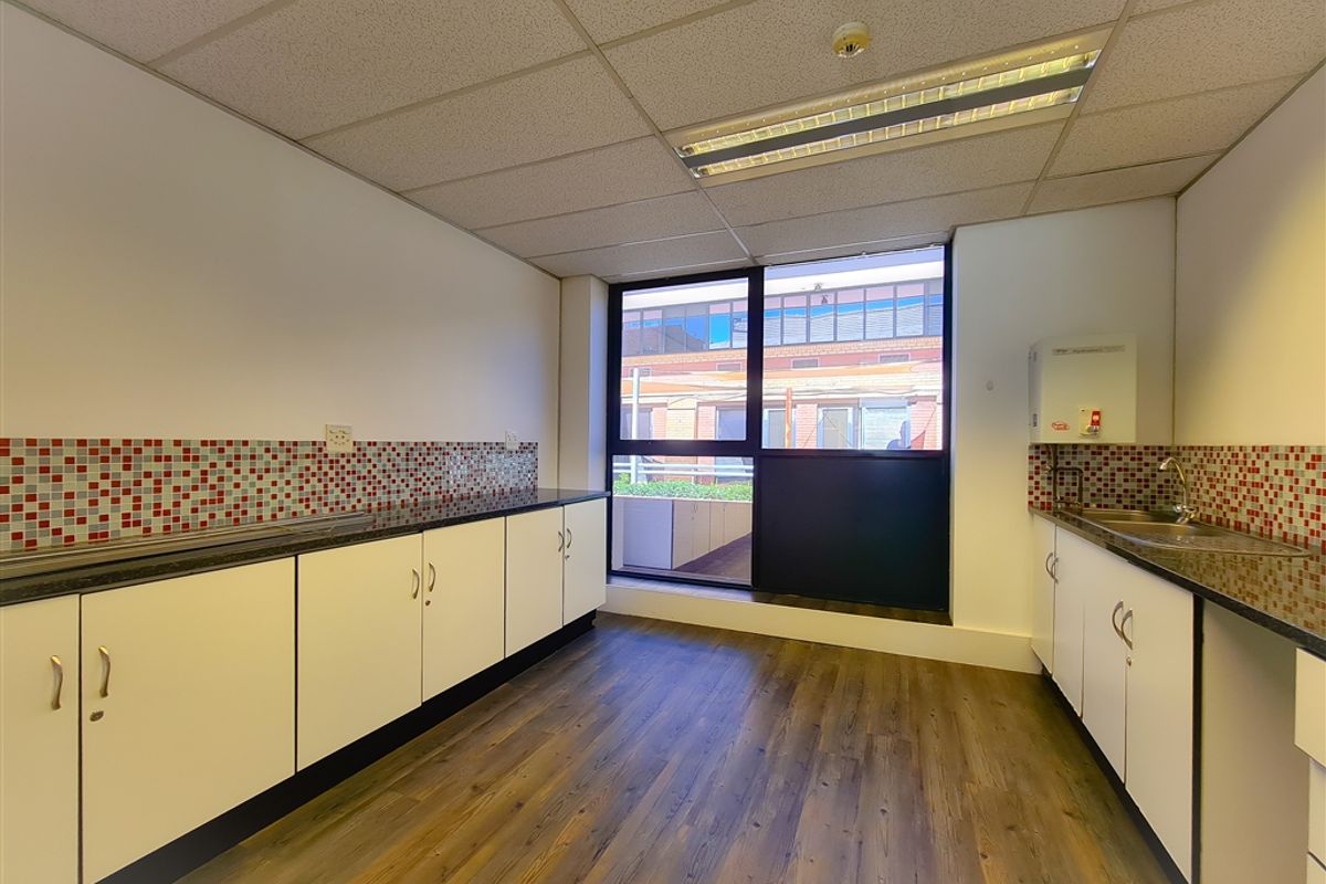 A functional office kitchenette featuring white cabinetry, dark countertops, mosaic tile backsplash, wood-look flooring, and a large window providing natural light.