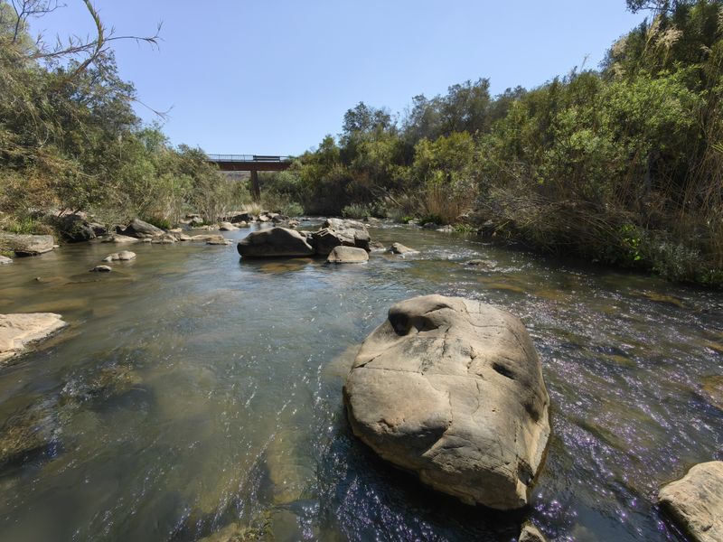 A scenic view of a natural river flowing through the farm landscape, featuring large rock formations and a bridge in the distance.