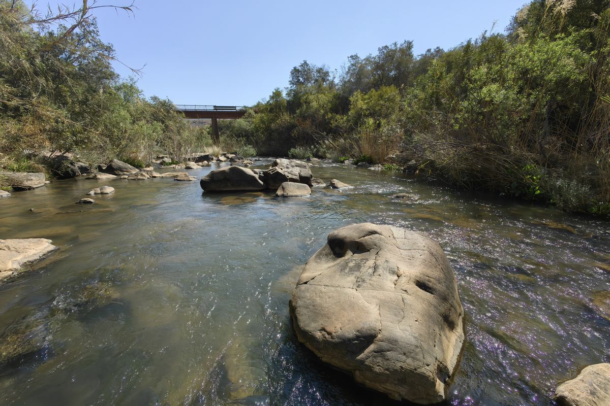 A scenic view of a natural river flowing through the farm landscape, featuring large rock formations and a bridge in the distance.