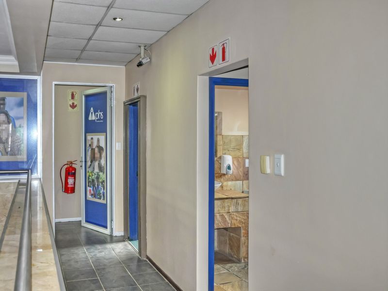 A functional office hallway featuring tiled flooring, a suspended ceiling with recessed lighting, and access to multiple rooms including a restroom area.