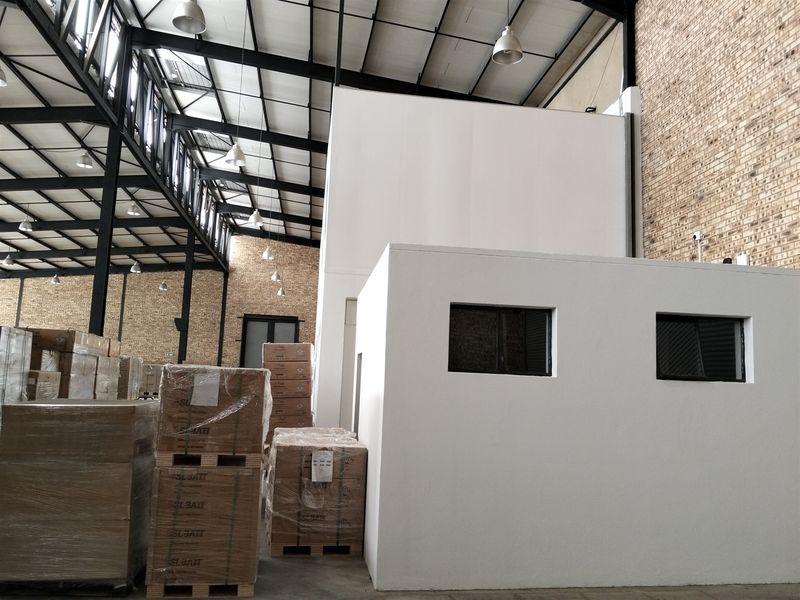 Spacious industrial warehouse interior featuring exposed steel roof trusses, brick wall finishes, and a built-in partitioned office structure.