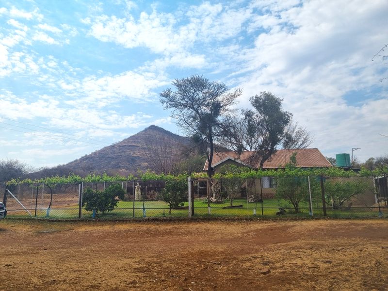 Front view of a farm property featuring a tiled roof dwelling, perimeter wire fencing with trellis, and a scenic mountain backdrop.
