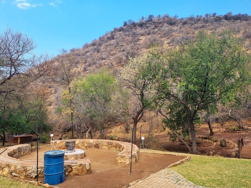 An outdoor garden area featuring a stone-clad fire pit, paved pathways, and natural landscape views against a hillside backdrop.