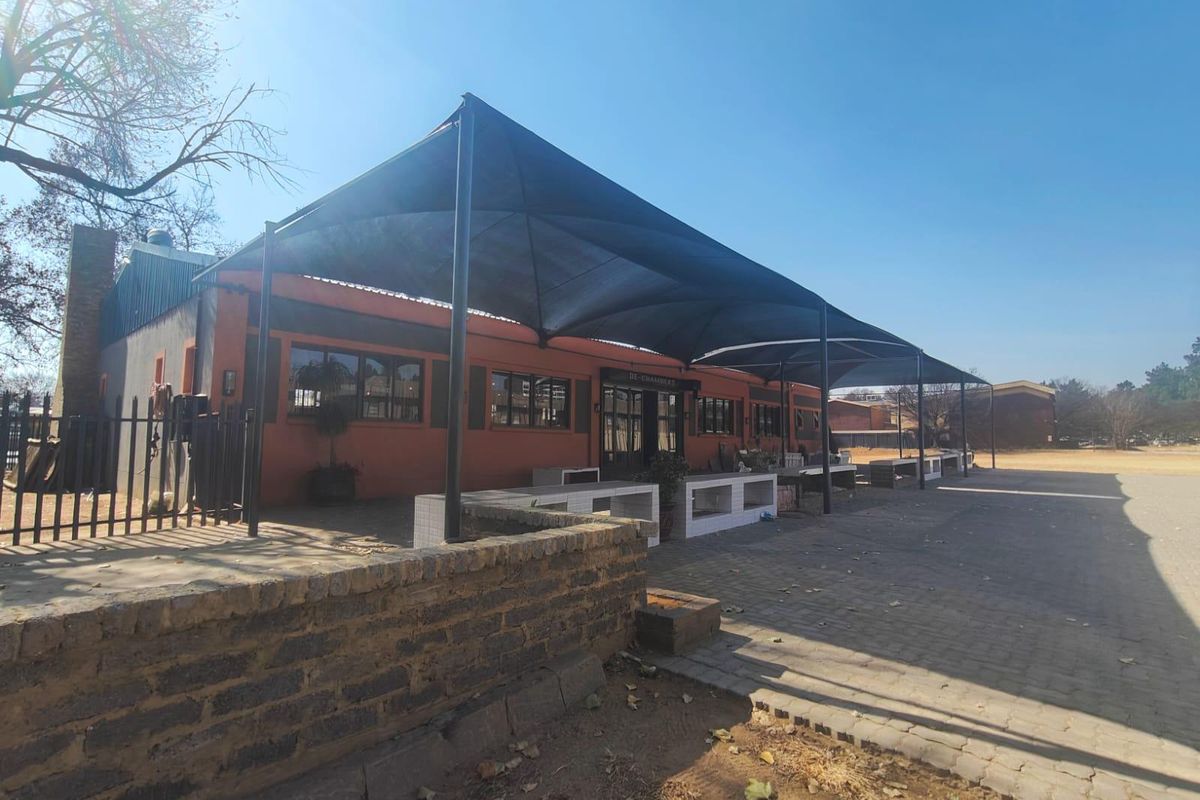 Exterior view of a commercial building featuring a large shade sail covering a paved patio area with built-in brick counters.