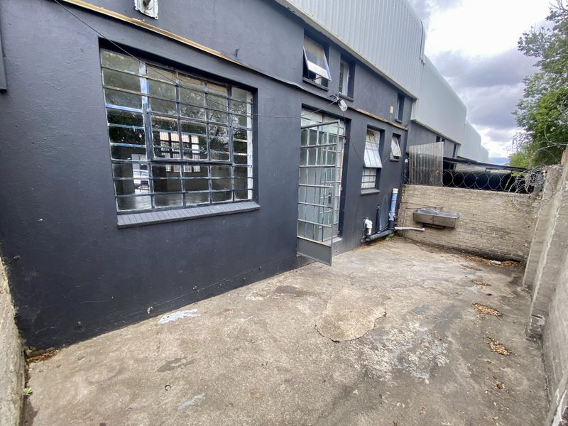Rear exterior area of an industrial property featuring concrete paving, a utility sink, and steel-framed windows and doors against a dark-painted facade.