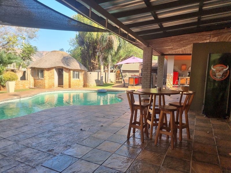 Covered outdoor patio area overlooking a swimming pool and a traditional thatched lapa, featuring tiled flooring and brick structural pillars.