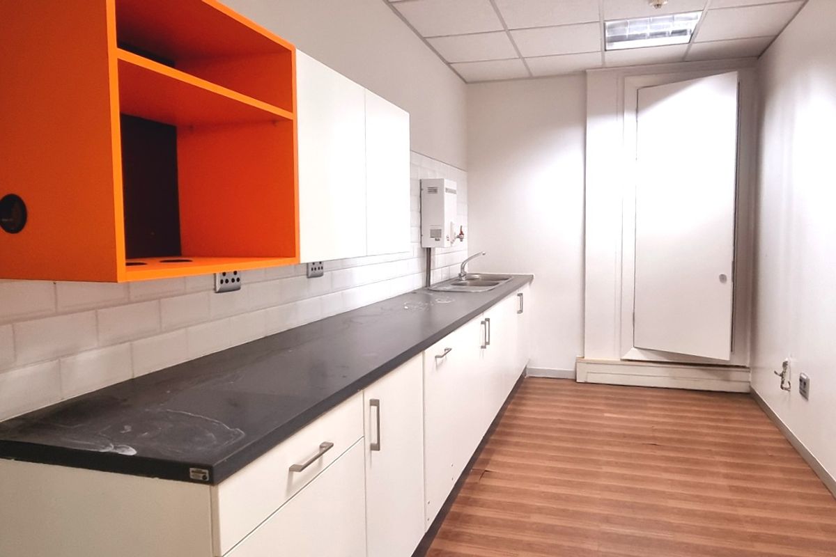 A functional office kitchenette featuring white cabinetry, a contrasting orange open shelf, a dark countertop, and a stainless steel sink.