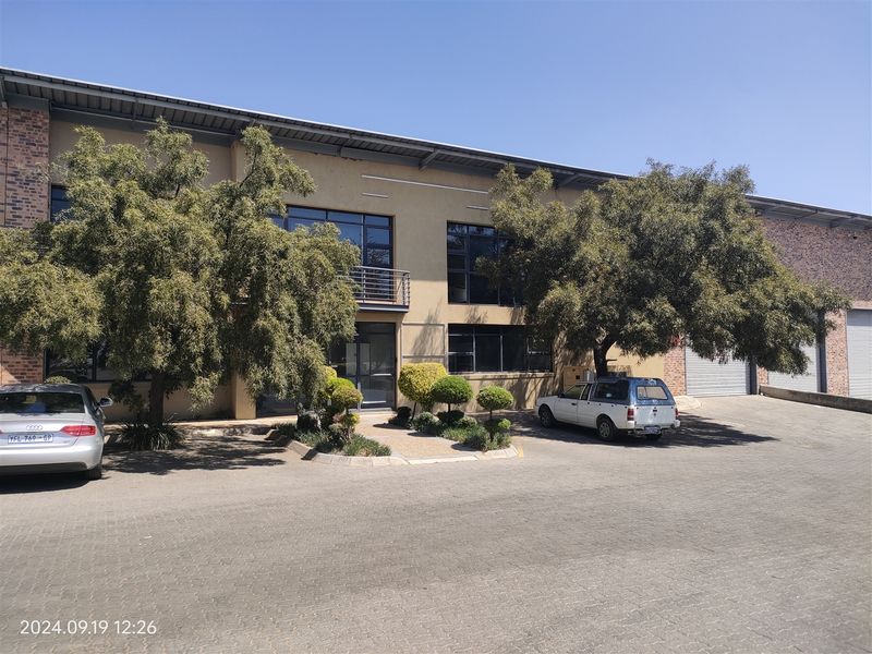 Front elevation of an industrial property featuring a brick and plaster facade, large windows, a small balcony, and paved driveway access with roller shutter doors.