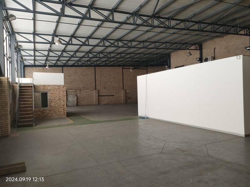 Spacious industrial warehouse featuring exposed brick walls, high steel truss ceilings, concrete flooring, and a built-in mezzanine staircase area.