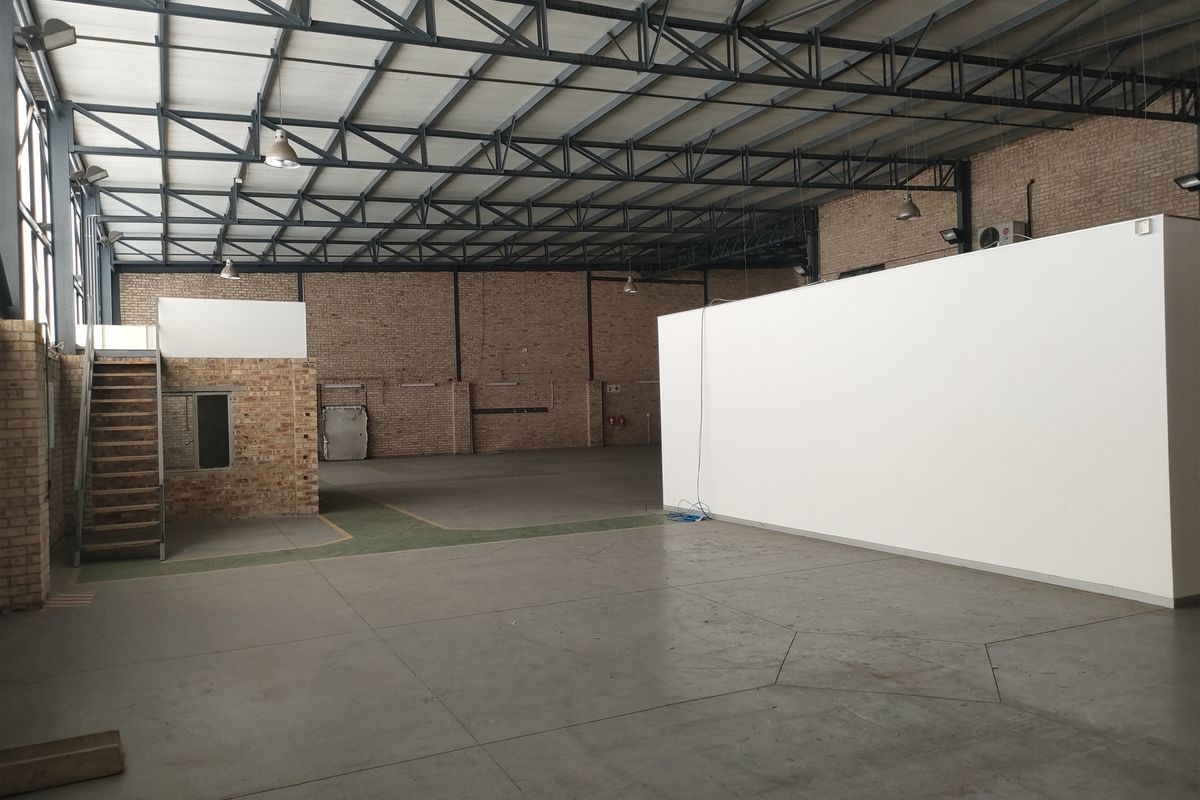 Spacious industrial warehouse featuring exposed brick walls, high steel truss ceilings, concrete flooring, and a built-in mezzanine staircase area.