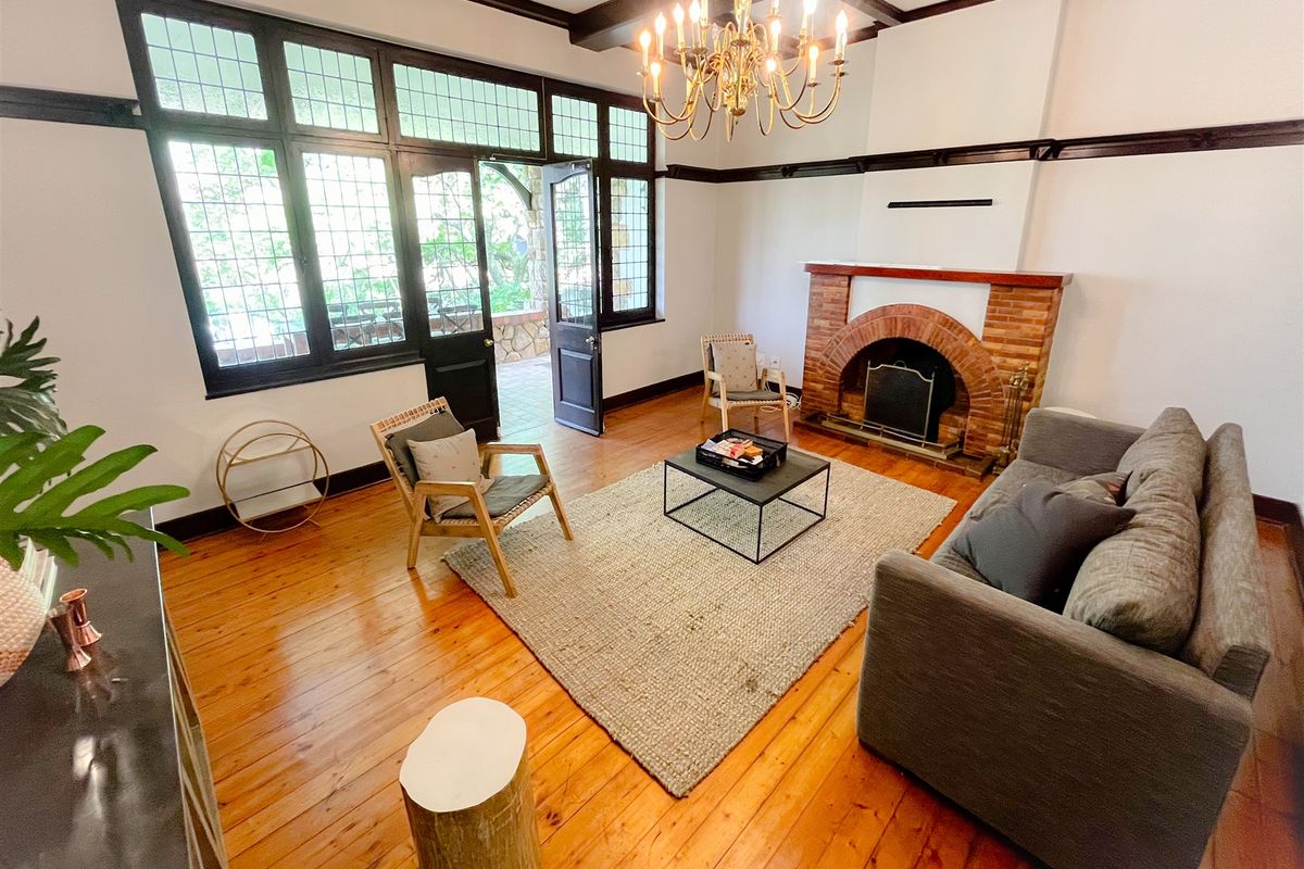 Spacious living room featuring original hardwood flooring, exposed timber ceiling beams, a classic brick fireplace, and large leaded glass windows with garden access.