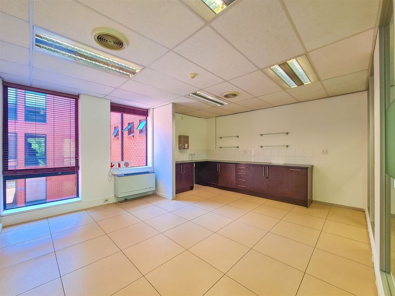 A bright, empty office space featuring tiled flooring, built-in cabinetry, suspended ceilings with recessed lighting, and large windows with venetian blinds.