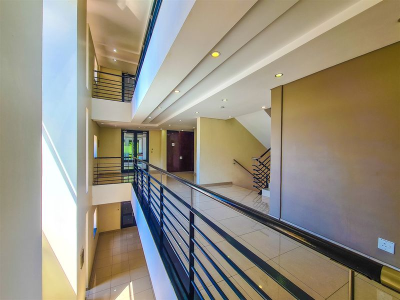 A bright, modern multi-level office hallway featuring tiled flooring, recessed lighting, and a sleek metal balustrade overlooking the atrium.