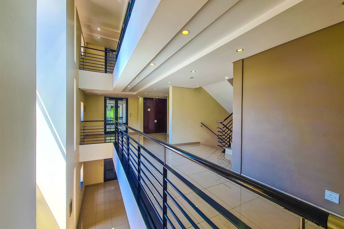 A bright, modern multi-level office hallway featuring tiled flooring, recessed lighting, and a sleek metal balustrade overlooking the atrium.