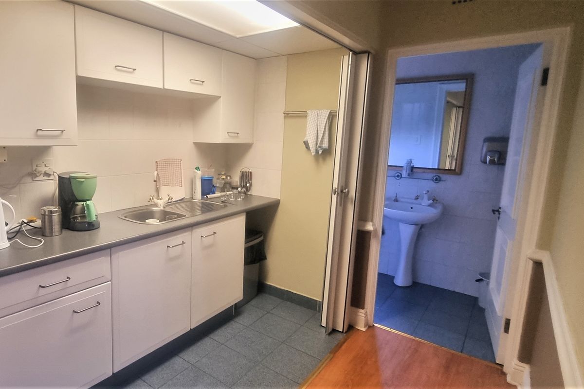 A functional office kitchenette featuring white cabinetry, a stainless steel sink, and tiled flooring, with an adjacent small bathroom.