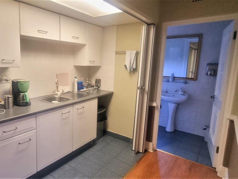 A functional office kitchenette featuring white cabinetry, a stainless steel sink, and tiled flooring, with an adjacent small bathroom.