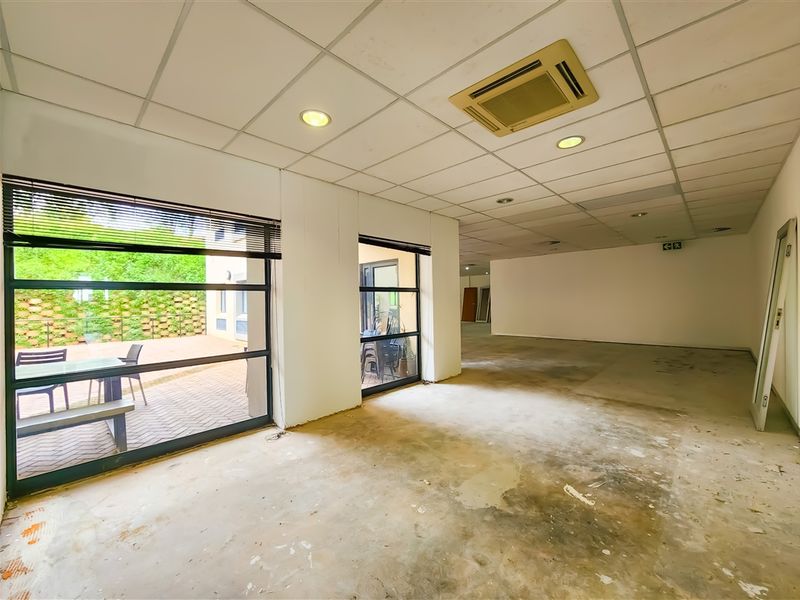 An empty, open-plan office space featuring a suspended ceiling, recessed lighting, integrated air conditioning, and large windows overlooking an outdoor patio area.